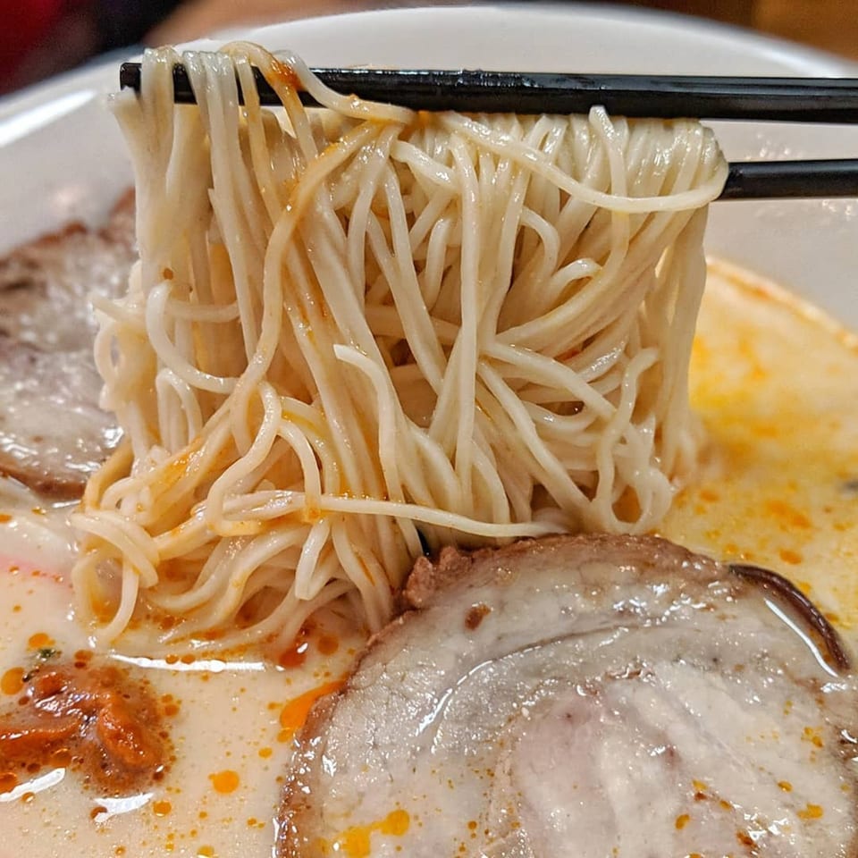 Chopsticks lifting a tangle of Momo Ramen noodles from a rich, creamy broth with pork slices.