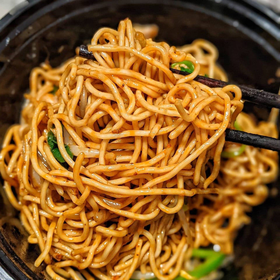 Chopsticks lifting a tangle of saucy ramen noodles with green onions from a black bowl.